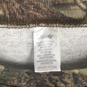 May include: Close-up of a camouflage-patterned sweatshirt with a white tag. The tag reads "Liberty" and includes size and material information: 80% cotton, 20% polyester. The sweatshirt is a 2X-Large regular size.