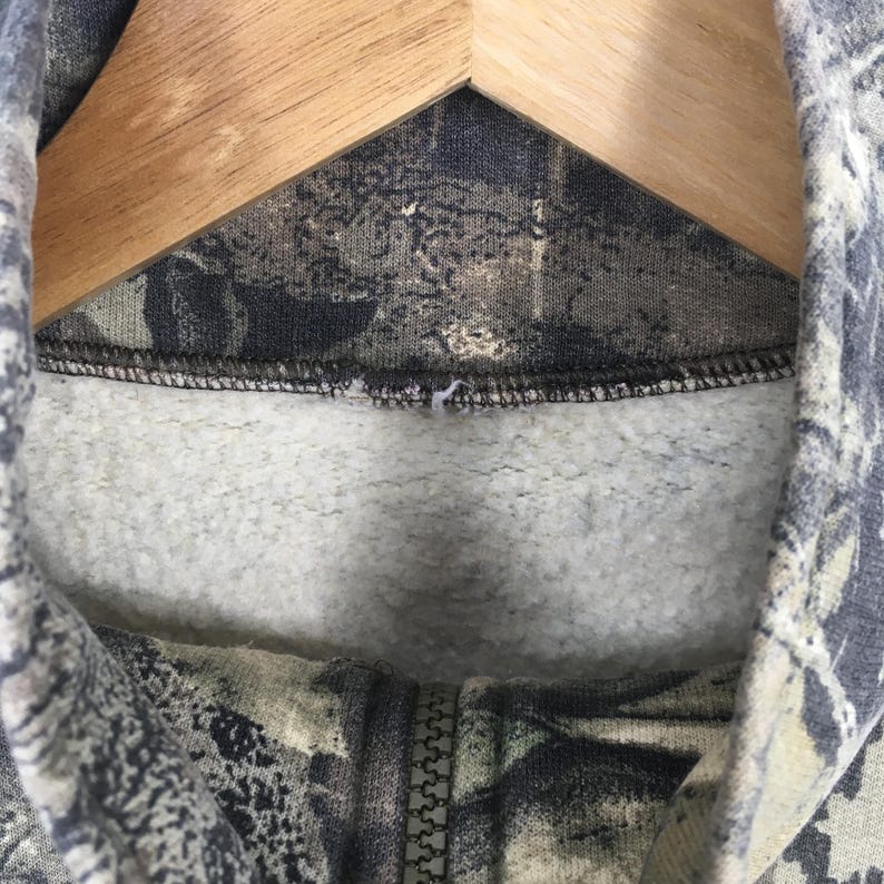 Pode incluir: Close-up de um moletom com capuz com padr&atilde;o de camuflagem. O tecido apresenta tons de verde, castanho e cinza. O z&iacute;per &eacute; vis&iacute;vel e o forro interior &eacute; cinza claro. O moletom est&aacute; pendurado em um cabide de madeira.