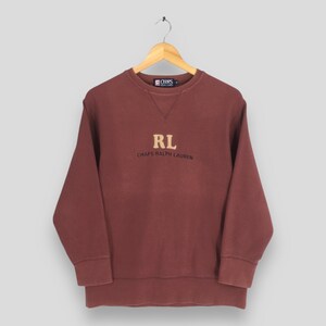 May include: A maroon long-sleeve crewneck sweatshirt featuring a tan embroidered RL logo and the text CHAPS RALPH LAUREN in black lettering on the chest. The garment includes a ribbed collar, cuffs, and hem, with a Chaps brand label at the neck.