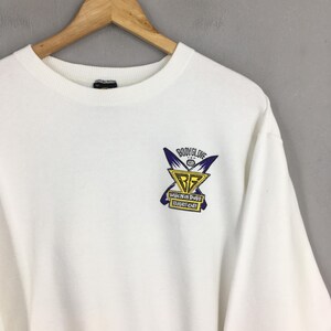 Op de afbeelding: Een witte crewneck sweatshirt met een klein, kleurrijk logo op de borst. Het logo bevat de woorden "Body Glove" boven een geel en blauw embleem met de tekst "When in Doubt Paddle Out!"