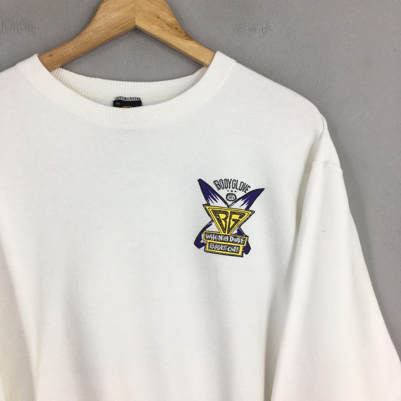 Op de afbeelding: Een witte crewneck sweatshirt met een klein, kleurrijk logo op de borst. Het logo bevat de woorden "Body Glove" boven een geel en blauw embleem met de tekst "When in Doubt Paddle Out!"