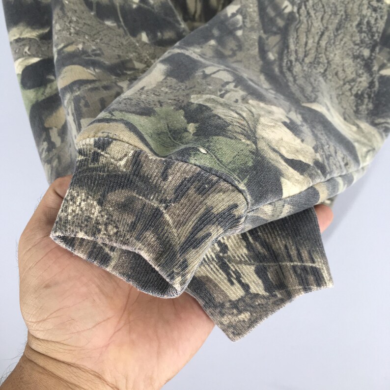 Pode incluir: Close-up de uma manga de camisola com padr&atilde;o de camuflagem. O tecido apresenta uma mistura de verdes, castanhos e castanhos claros, criando um efeito de floresta. O punho &eacute; canelado e mostra desgaste.