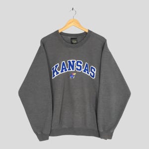 May include: A dark grey sweatshirt featuring the word "KANSAS" in blue, outlined in white, across the chest. A small Jayhawk graphic is beneath the text. The sweatshirt is suspended from a wooden hanger.