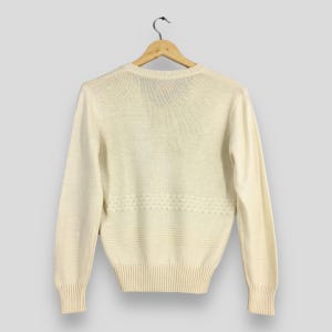 May include: Cream-colored sweater with long sleeves, ribbed cuffs, and a ribbed hem. The sweater features a decorative horizontal pattern across the midsection. The garment is hanging on a wooden hanger against a neutral background.
