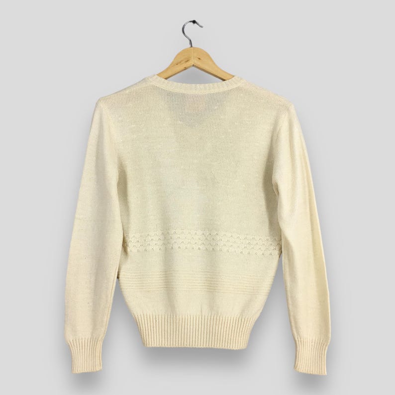 May include: Cream-colored sweater with long sleeves, ribbed cuffs, and a ribbed hem. The sweater features a decorative horizontal pattern across the midsection. The garment is hanging on a wooden hanger against a neutral background.
