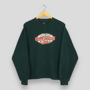 Puede incluir: Una sudadera verde oscuro con cuello redondo y mangas largas. La parte delantera presenta un gráfico con el texto "Atlantic League Champions 20 DUCKS 04" en rojo, naranja y blanco. La sudadera está colgada en una percha de madera.