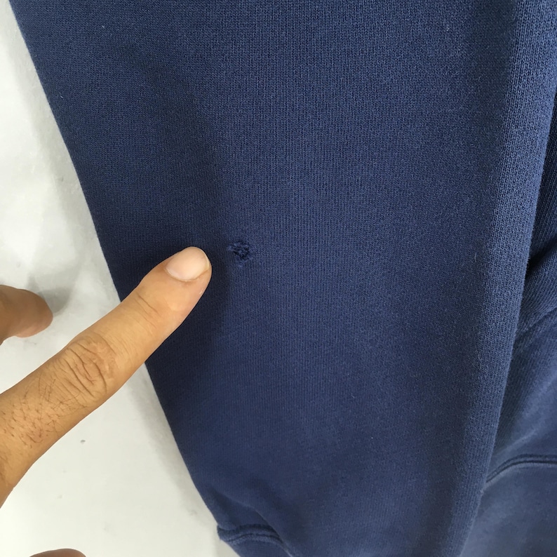 Pu&ograve; includere: Primo piano di una manica di felpa blu navy con un piccolo foro. Il tessuto sembra essere un materiale morbido e lavorato a maglia. Un dito indica il foro nella manica.