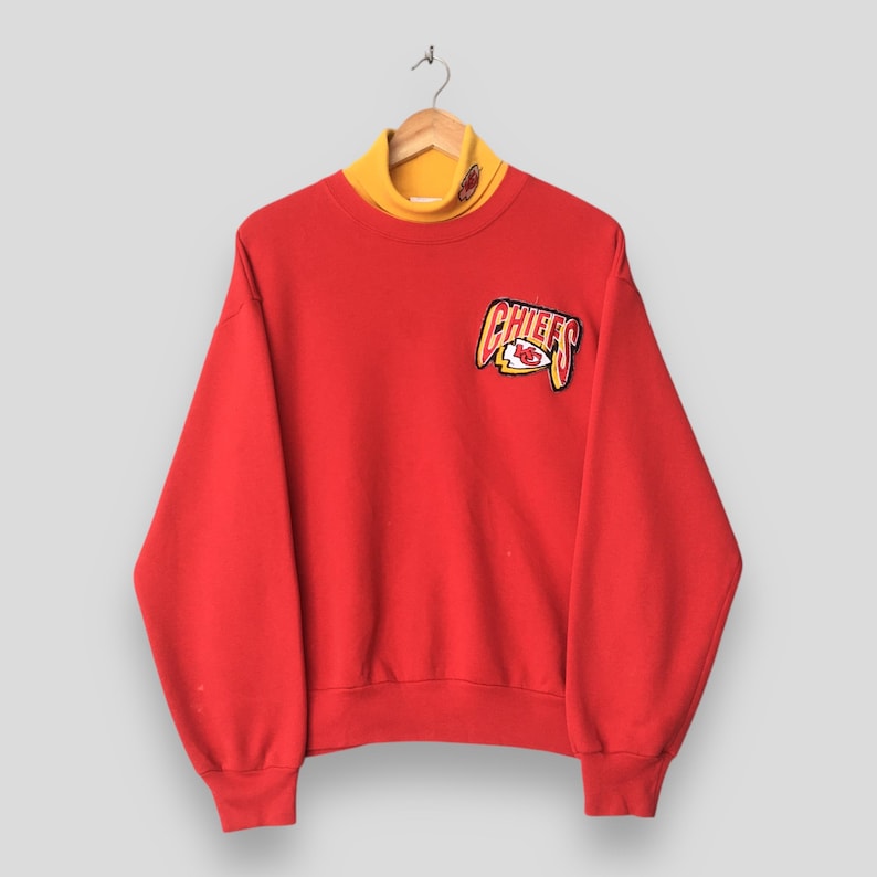 Peut inclure: Un sweat-shirt rouge &agrave; manches longues avec un col montant jaune et un &eacute;cusson Kansas City Chiefs sur la poitrine droite. Le logo affiche le mot CHIEFS en lettres jaunes et rouges accompagn&eacute; d'un embl&egrave;me en forme de pointe de fl&egrave;che.