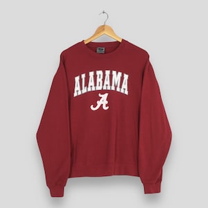 May include: A maroon sweatshirt with the word "ALABAMA" in white block letters and a white "A" logo. The sweatshirt is hanging on a wooden hanger against a neutral background. The long-sleeved top is made of a soft material.