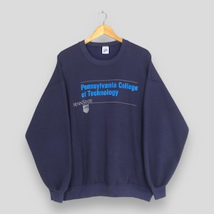 Puede incluir: Sudadera azul marino con el texto "Pennsylvania College of Technology" en azul y blanco. La sudadera también presenta el logotipo de Penn State. La sudadera está colgada en una percha de madera.