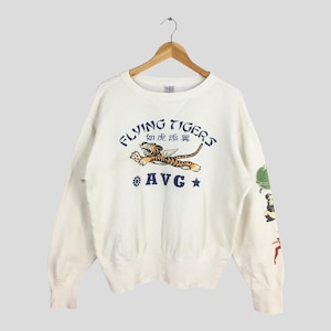 Puede incluir: Sudadera de color crema con el texto "Flying Tigers" y un gráfico de tigre. El diseño incluye el texto "AVG" y una estrella. Gráficos adicionales en la manga derecha.