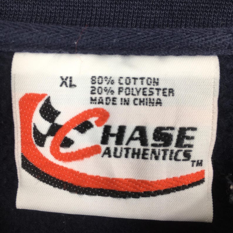 Puede incluir: Una etiqueta blanca con el texto "XL", "80% COTTON 20% POLYESTER MADE IN CHINA". La etiqueta presenta un logotipo rojo y negro y las palabras "CHASE AUTHENTICS". La etiqueta est&aacute; adherida a una prenda azul marino.