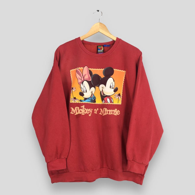 May include: A red sweatshirt featuring a graphic of Mickey and Minnie Mouse. The graphic is in a rectangular frame with a yellow and orange background. The words "Mickey n' Minnie" are printed below the image. The sweatshirt is hanging on a wooden hanger.