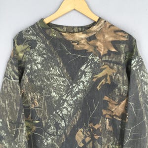 Puede incluir: Sudadera con estampado de camuflaje y cuello redondo, en tonos marrones, verdes y grises. El dise&ntilde;o imita un entorno forestal, con detalles de corteza de &aacute;rbol y hojas. La parte superior de manga larga est&aacute; colgada en una percha de madera.