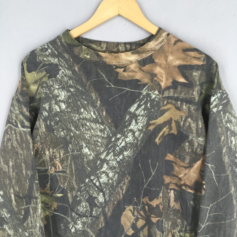 Puede incluir: Sudadera con estampado de camuflaje y cuello redondo, en tonos marrones, verdes y grises. El dise&ntilde;o imita un entorno forestal, con detalles de corteza de &aacute;rbol y hojas. La parte superior de manga larga est&aacute; colgada en una percha de madera.