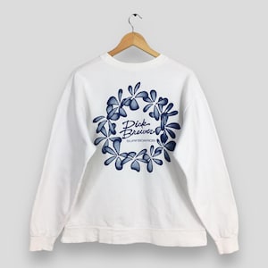 Puede incluir: Sudadera blanca con un diseño floral azul y el texto "Dick Brewer SURFBOARDS" en el centro. La sudadera está colgada en una percha de madera.