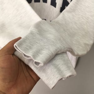 May include: Close-up of a gray and white sweatshirt sleeve. The cuff is a darker gray, ribbed knit. The body of the sweatshirt is a lighter gray. The top of the sweatshirt has dark blue lettering.