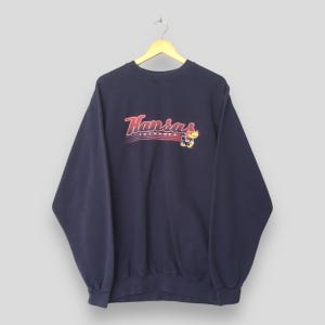 May include: Navy blue sweatshirt featuring the word "Kansas" in red script. Below the word "Kansas" is the word "Jayhawks". A cartoon Jayhawk mascot is on the right. The sweatshirt is hanging from a wooden hanger.