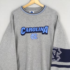 Puede incluir: Una sudadera gris con la palabra "CAROLINA" en letras azules claras en el pecho. Debajo, un logotipo azul claro y azul marino. El cuello tiene rayas azul claro y azul marino. Una franja azul marino con un logotipo blanco en la manga.