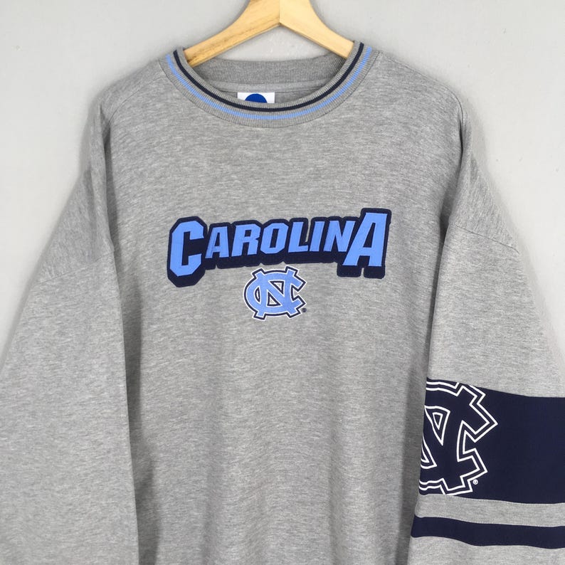 Puede incluir: Una sudadera gris con la palabra "CAROLINA" en letras azules claras en el pecho. Debajo, un logotipo azul claro y azul marino. El cuello tiene rayas azul claro y azul marino. Una franja azul marino con un logotipo blanco en la manga.