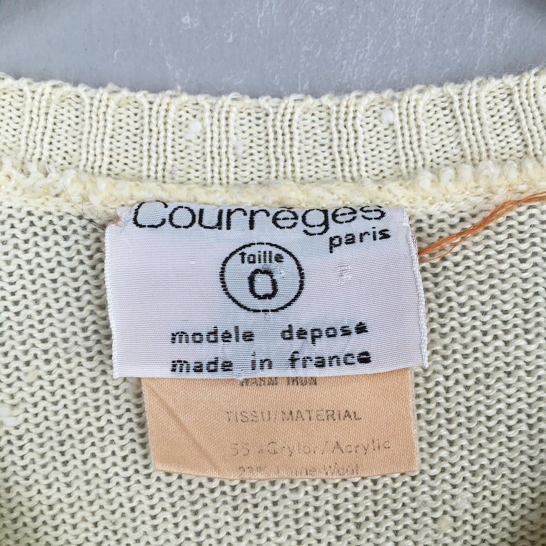 May include: Close-up of a cream-colored knit garment with a label. The label reads "Courreges paris" and "modele depose made in france." The label also indicates the size is "0" and the material is 55% Grylor/Acrylic.