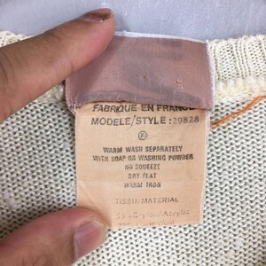 May include: Close-up of a cream-colored knit fabric with a sewn-in tag. The tag reads "FABRIQUE EN FRANCE" and includes washing instructions. The fabric is a blend of Grylor/Acrylic and Laine Wool.