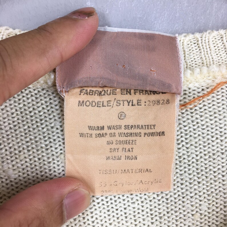 May include: Close-up of a cream-colored knit fabric with a sewn-in tag. The tag reads "FABRIQUE EN FRANCE" and includes washing instructions. The fabric is a blend of Grylor/Acrylic and Laine Wool.