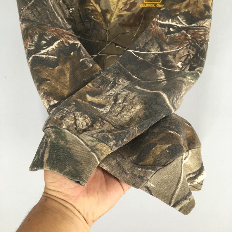 May include: A camouflage-patterned sweatshirt with brown, green, and tan hues. The sleeves are cuffed at the wrists. The brand name "KILLBUCK, OHIO" is visible on the upper part of the sweatshirt.