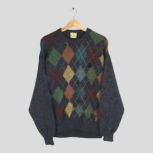 May include: A dark grey argyle sweater with a crew neck and long sleeves. The argyle pattern features diamonds in shades of green, brown, yellow, and blue. A small, dark emblem is on the chest. The sweater is hanging on a wooden hanger.