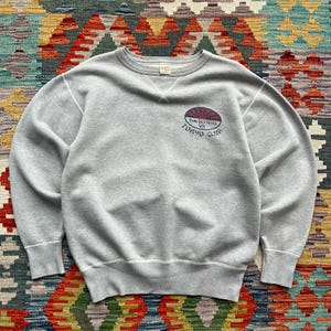 May include: A light gray crewneck sweatshirt featuring a ribbed collar with a V-shaped stitch detail. On the upper right chest, there is a graphic logo with a red and blue sun design, the text Sun Setters VII, and the words Tokyo Club below it.