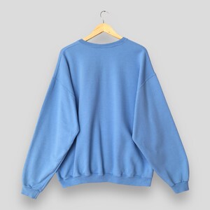 May include: A light blue sweatshirt with a crew neck and long sleeves, hanging on a wooden hanger. The sweatshirt is a solid color and appears to be made of a soft material. The back of the sweatshirt is visible.
