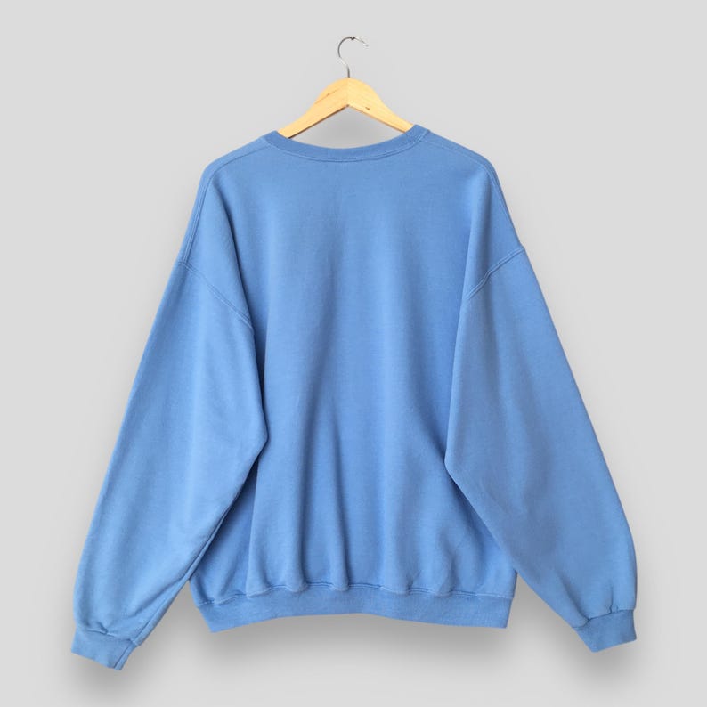 May include: A light blue sweatshirt with a crew neck and long sleeves, hanging on a wooden hanger. The sweatshirt is a solid color and appears to be made of a soft material. The back of the sweatshirt is visible.