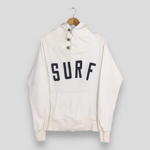 May include: A white hoodie with the word "SURF" in navy blue block letters across the chest. The hoodie has a unique collar with three buttons and a front pocket. The sleeves and bottom hem have ribbed cuffs.