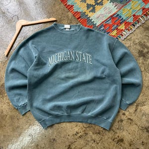 Puede incluir: Una sudadera de cuello redondo de color verde azulado con el texto bordado "MICHIGAN STATE" en blanco. La sudadera está tendida sobre una superficie texturizada, con una percha de madera y una alfombra colorida en el fondo.