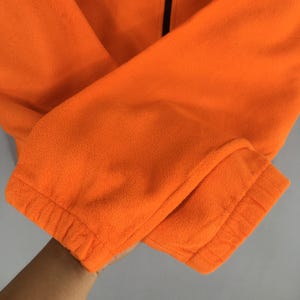May include: Close-up of an orange fleece jacket sleeve. The sleeve is folded, showing the elastic cuff and the soft texture of the fleece material. A black zip is visible.