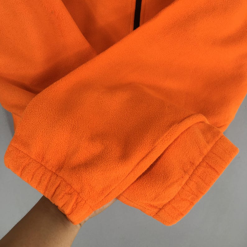 May include: Close-up of an orange fleece jacket sleeve. The sleeve is folded, showing the elastic cuff and the soft texture of the fleece material. A black zip is visible.