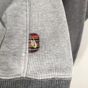 K&ouml;nnte beinhalten: Nahaufnahme eines grauen Sweatshirts mit gerippter Textur. Ein kleines, rechteckiges Patch mit dem Nike-Logo ist auf dem &Auml;rmel aufgen&auml;ht. Das Patch hat ein farbenfrohes Streifendesign und den Nike-Swoosh.