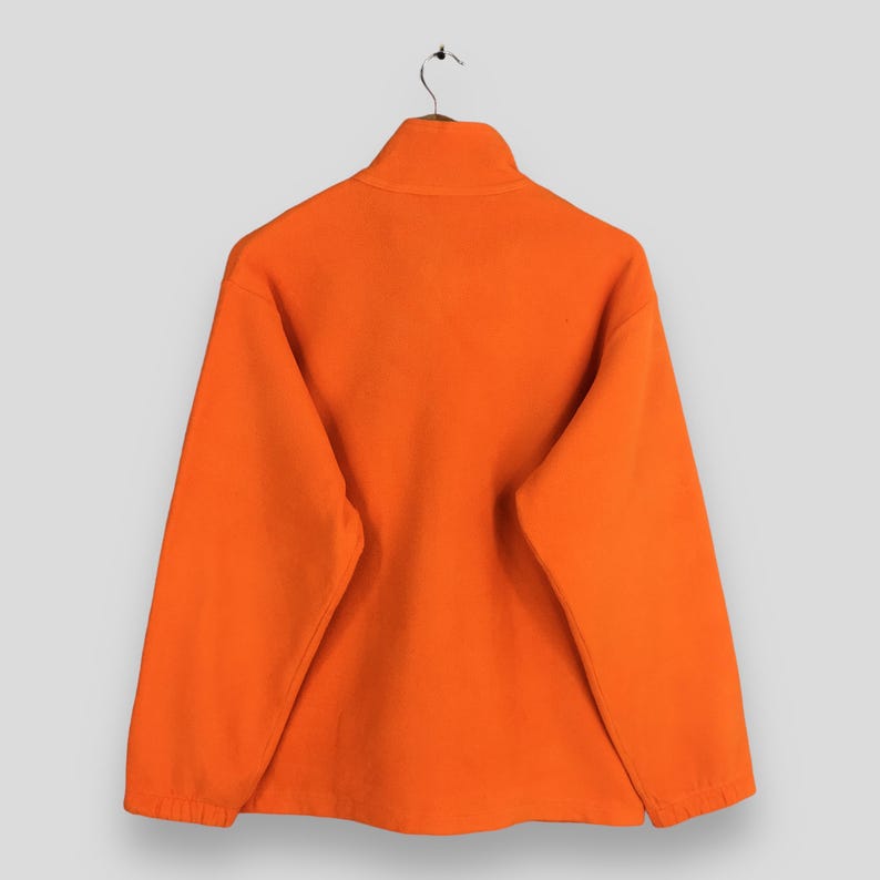 May include: Vibrant orange fleece pullover with a high collar and long sleeves, hanging on a metal hanger. The material looks soft and warm, ideal for cooler temperatures. The garment is displayed against a plain background.