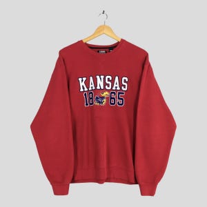 May include: A red crewneck sweatshirt with the word "KANSAS" in white block letters, the numbers "18" and "65", and a Jayhawk mascot graphic. The sweatshirt is hanging on a wooden hanger.