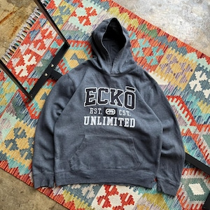 May include: A charcoal gray hoodie with the text "ECKO UNLIMITED" in white block letters. The hoodie has a front pocket and a hood. The hoodie is lying on a colorful patterned rug.