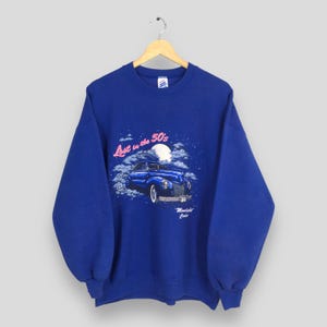 Puede incluir: Una sudadera azul real con un gráfico de un coche vintage y el texto "Lost in the 50's" en rosa. El coche es azul y tiene una luna blanca y nubes en el fondo.