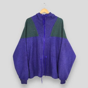 Könnte beinhalten: Eine lila-grüne Fleecejacke mit durchgehendem Reißverschluss und Kapuze. Die Jacke hat einen Kordelzug in der Taille und an den Manschetten. Das Columbia-Logo ist auf der Brust sichtbar. Hängt an einem Holzkleiderbügel.