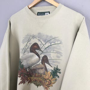 Peut inclure: Un sweat-shirt beige avec un imprim&eacute; de deux canards dans un d&eacute;cor d'automne. Le texte "The Sportsman" est imprim&eacute; sur le sweat-shirt.
