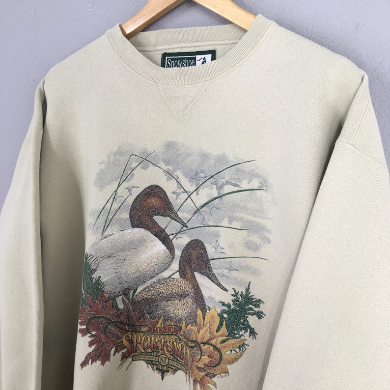 Peut inclure: Un sweat-shirt beige avec un imprim&eacute; de deux canards dans un d&eacute;cor d'automne. Le texte "The Sportsman" est imprim&eacute; sur le sweat-shirt.