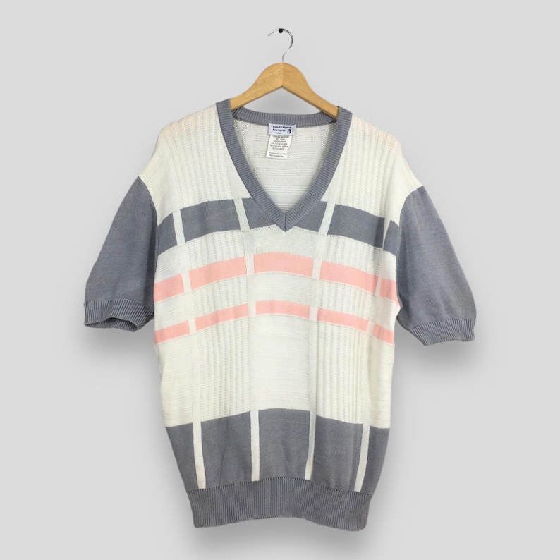 May include: A vintage short-sleeved sweater with a V-neck design. The sweater features a white base with gray and pink geometric patterns across the front. The sleeves and neckline are gray. The sweater is hanging on a wooden hanger.
