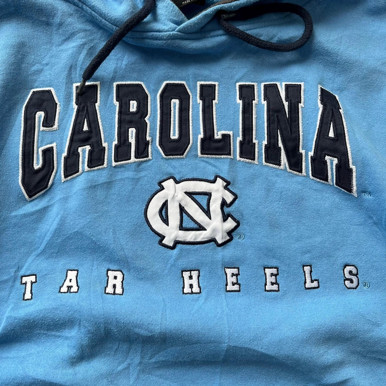 K&ouml;nnte beinhalten: Hellblauer Kapuzenpullover mit dem Wort "CAROLINA" in marineblauen Blockbuchstaben. Unter dem Wort "CAROLINA" befindet sich ein wei&szlig;es und marineblaues Logo. Die Worte "TAR HEELS" sind in marineblauen Blockbuchstaben gedruckt. Der Hoodie hat einen Kordelzug.