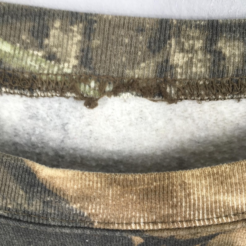 Puede incluir: Primer plano del cuello de una sudadera con estampado de camuflaje. La tela presenta tonos marrones, verdes y beige, con un interior blanco y afelpado visible en el escote. El cuello es acanalado y muestra signos de desgaste.