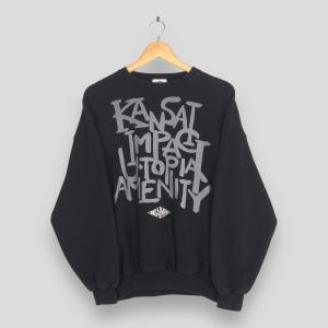 May include: Black crewneck sweatshirt with the words "KANSAS IMPACT UTOPIA AMENITY" in a gray, graffiti-style font. The sweatshirt is hanging on a wooden hanger against a light background. A small diamond-shaped logo is at the bottom.
