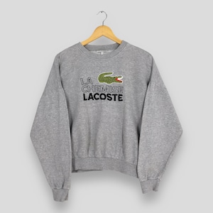 Może przedstawiać: Szara bluza z napisem "LA CHEMISE LACOSTE" w kolorze czarnym i białym oraz zielonym logo krokodyla z czerwonymi akcentami. Bluza wisi na drewnianym wieszaku.