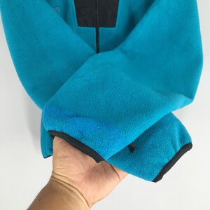May include: A turquoise fleece jacket with a black panel at the top and a black zipper. The cuffs are trimmed with black fabric. The jacket is held up by a hand.